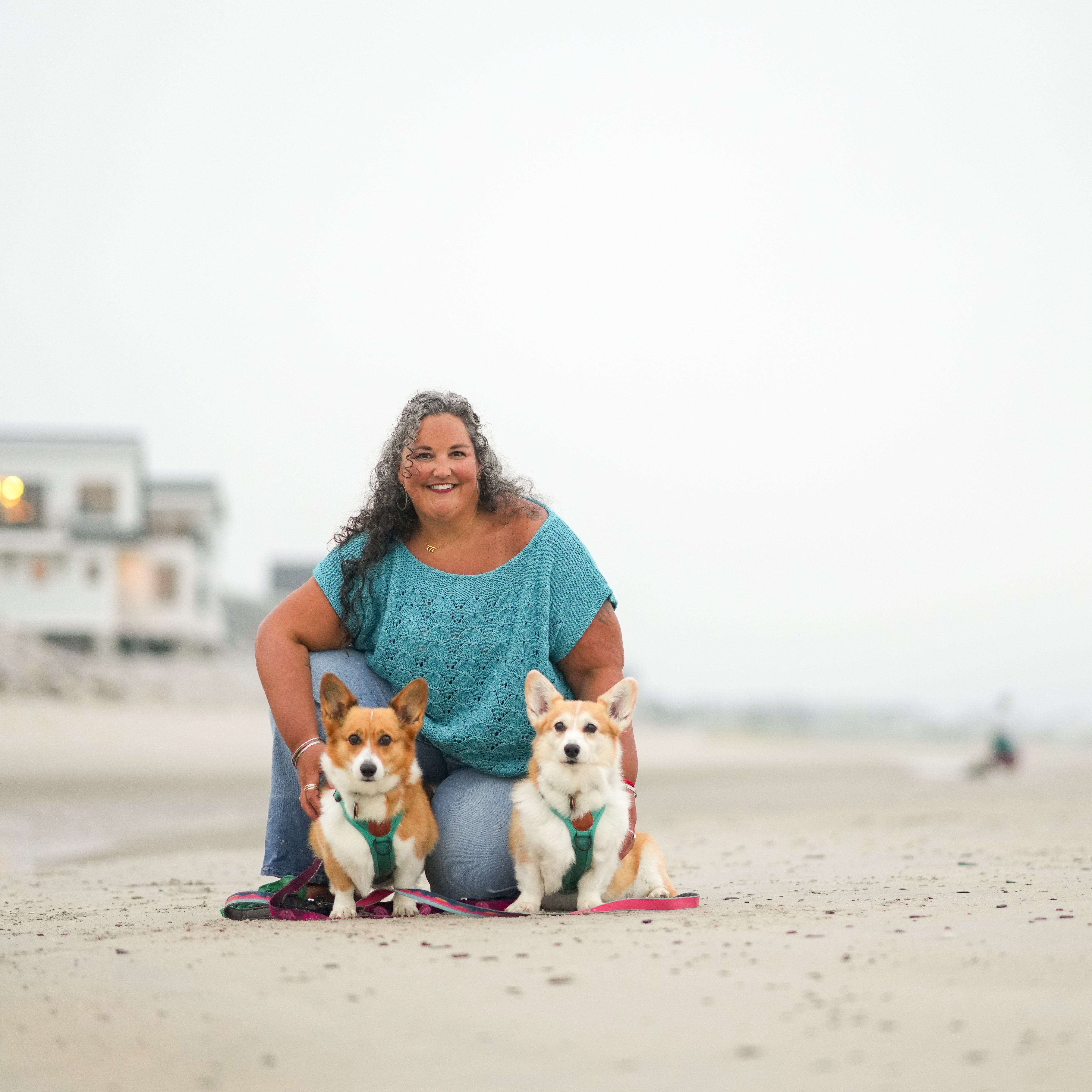 A 40 year old woman with graying long curly hair kneels on a beach, posing with her two corgis. 